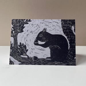May include: A black and white linocut print of a squirrel sitting on a tree branch, holding a nut. The print features a detailed illustration of the squirrel, oak tree, and acorns. The background is a light gray color.