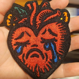May include: Embroidered patch of a red anatomical heart with a black border. The heart features a crying face with blue tears. The patch has orange, green, and yellow accents.