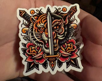 May include: A sticker featuring a traditional tattoo style illustration of a tiger and a panther head with a sword through the middle. The tiger and panther are both snarling with sharp teeth and the sword is silver with a black handle. The background is a red and green floral design.