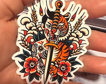 May include: A sticker featuring a traditional Japanese tattoo style illustration of a tiger with a sword through its head. The tiger is orange and black with white stripes. The sword is black with a gold handle. The background is white with blue and green floral elements.