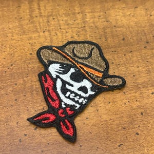 May include: An embroidered patch featuring a white skull wearing a brown cowboy hat with an orange band and a red bandana tied around the neck. The design is outlined in black thread, creating a distinct graphic character with a western theme.