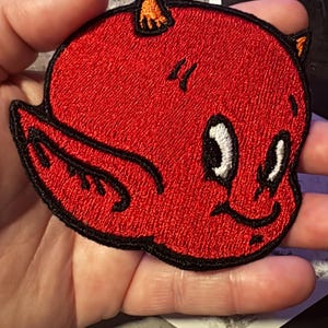 May include: A red embroidered patch featuring a cartoon devil with a smiling face, white eyes, and orange horns. The patch has a black border and is likely for clothing or accessories.