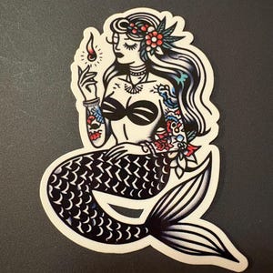 Puede incluir: Una pegatina de estilo tatuaje en blanco y negro de una sirena con un top de bikini negro, una corona floral y una cerilla encendida en la mano. Su cola es negra con escamas blancas.
