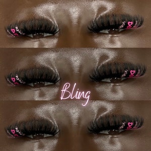 Könnte beinhalten: Nahaufnahme von Augen einer Person mit langen, dunklen Wimpern und rosafarbenem herzförmigem Glitzer an den äußeren Ecken. Das Wort "Bling" ist in rosa Buchstaben unter den Augen geschrieben.