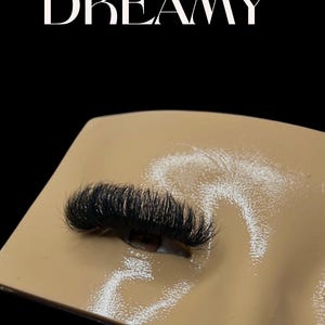 Könnte beinhalten: Nahaufnahme eines Paares künstlicher Wimpern auf einem beigen Wimpern-Übungspad. Die Wimpern sind lang und dick mit einer natürlichen Wölbung.