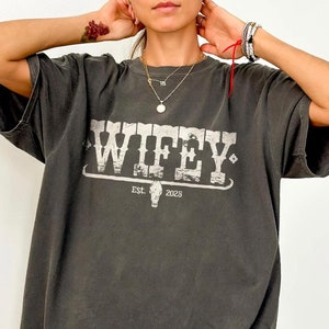 Könnte beinhalten: Ein überdimensioniertes T-Shirt in dunkelgrau mit einer weißen Grafik, auf der "WIFEY" mit einem Cowboyschädel und "EST. 2025" darunter steht.