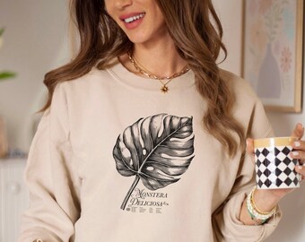 May include: A beige sweatshirt with a black and white illustration of a Monstera Deliciosa plant. The text "Monstera Deliciosa" is printed below the plant.