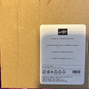 Puede incluir: Una etiqueta rectangular blanca con texto negro, adherida a un respaldo de cartón marrón. La etiqueta dice "STAMPIN' BLENDS STORAGE" e incluye texto en francés, alemán y japonés. Dimensiones: 12,7 x 12,7 cm.