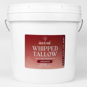 Op de afbeelding: Een witte emmer met een rood en wit etiket. Het etiket luidt "HAUS OF SAGE WHIPPED TALLOW FACE + BODY MOISTURIZER UNSCENTED NET 6.35 KG MADE IN THE USA 100% GRASS-FED BEEF". De emmer heeft een metalen handvat.