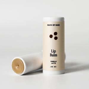 May include: Two white lip balm tubes, one lying down, the other upright. The upright tube has the text "HAUS OF SAGE" and "Lip Balm VANILLA LATTE .20 OZ". The exposed balm is light brown.