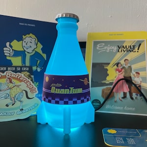 May include: A blue glowing light-up bottle shaped like a rocket with a label that reads "Nuka Cola QuanTum". The bottle is sitting on a table with other Fallout themed items.