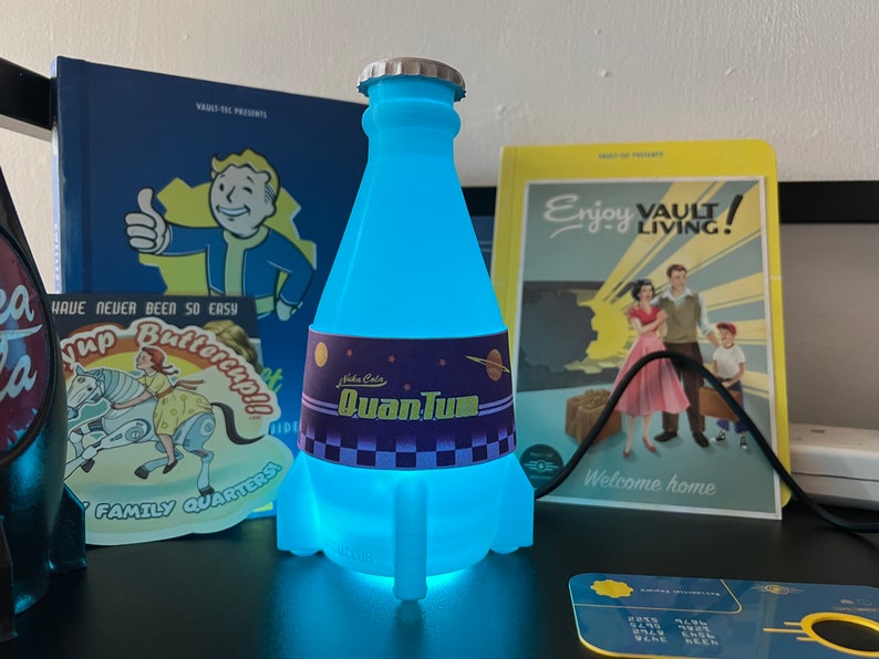 May include: A blue glowing light-up bottle shaped like a rocket with a label that reads "Nuka Cola QuanTum". The bottle is sitting on a table with other Fallout themed items.