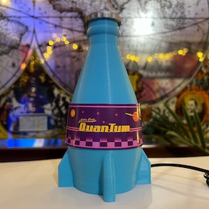 May include: A blue rocket-shaped bottle with a label that reads "Nukla Cola Quantum". The bottle has a silver cap and a purple and pink label with stars and a planet.