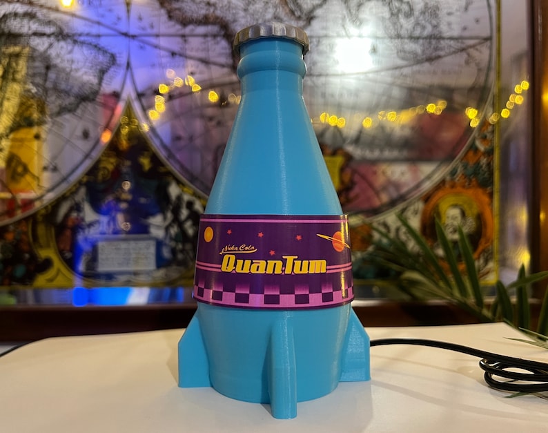 May include: A blue rocket-shaped bottle with a label that reads "Nukla Cola Quantum". The bottle has a silver cap and a purple and pink label with stars and a planet.
