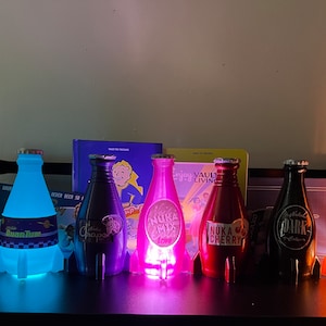 May include: Seven different colored glass bottles with labels and rocket-shaped bases. The bottles are lit from within, casting a glow. The labels include "Nuka Cola", "Quantum", "Nuka Mix", "Nuka Cherry", "Dark", and "Victory".