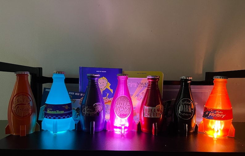 May include: Seven different colored glass bottles with labels and rocket-shaped bases. The bottles are lit from within, casting a glow. The labels include "Nuka Cola", "Quantum", "Nuka Mix", "Nuka Cherry", "Dark", and "Victory".