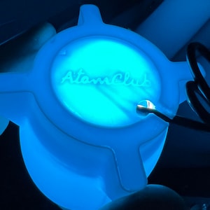 May include: A blue plastic light housing with the text "Atom Club" printed on the top. The housing is designed to fit on the bottom lid of a product.