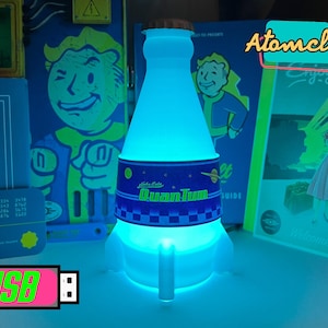 May include: A blue and white light-up bottle shaped like a rocket with the text "Nuka Cola" and "Quan Tum" on the label. The bottle is lit from within, creating a blue glow. The bottle is a USB-powered novelty lamp.