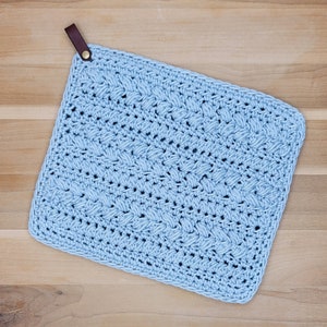 Pode incluir: Um pano de prato de crochê azul claro com uma alça de couro marrom para pendurar.