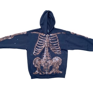 Peut inclure: Sweat à capuche bleu marine avec une impression de squelette en beige et marron. Le motif squelettique recouvre le devant, le dos et les manches. Le sweat à capuche est zippé et possède une capuche, idéal pour un thème d'Halloween.