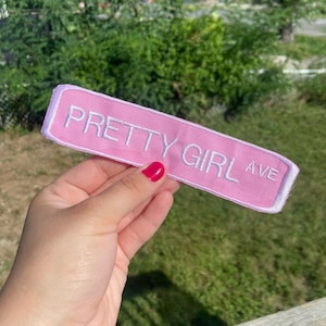 Op de afbeelding: Een roze geborduurde patch met de woorden "PRETTY GIRL AVE" in het wit. De patch heeft een witte rand en is rechthoekig van vorm. De patch wordt voor een groene achtergrond gehouden.