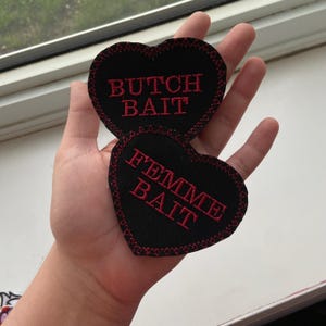May include: Two black felt heart-shaped patches with red embroidered text. The top patch reads "Butch Bait" and the bottom patch reads "Femme Bait".