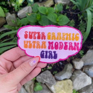 May include: A pink, cloud-shaped patch with the text "Super Graphic Ultra Modern Girl" in a retro font. The text is in different colors, including orange, yellow, red, and purple.