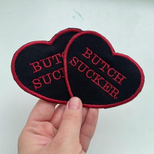 Puede incluir: Dos parches negros en forma de corazón con texto bordado en rojo "BUTCH SUCKER". Los parches tienen un borde rojo. Los parches están sostenidos por una mano.