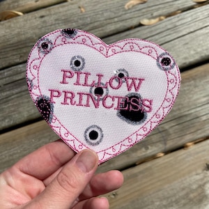 Op de afbeelding: Een hartvormige patch met de woorden "PILLOW PRINCESS" geborduurd in roze. De patch heeft een roze rand en is versierd met zwarte en zilveren accenten. De patch staat op een witte achtergrond.