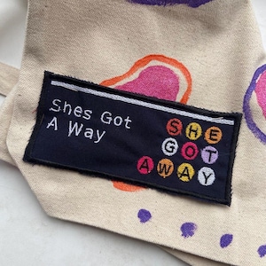 May include: A black fabric patch with white embroidery that reads "She's Got A Way" and a colourful circle design that reads "SHE GOT AWAY".