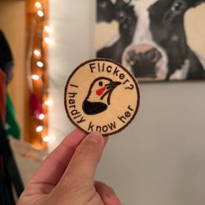 May include: A circular embroidered patch with a beige background and a dark brown border. It features a stylized bird head with a red cheek and black throat, surrounded by the black embroidered text "Flicker? I hardly know her."