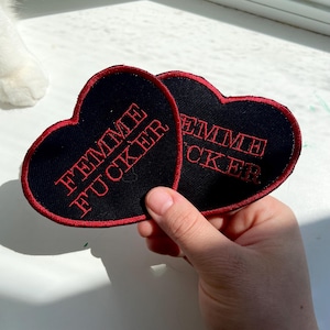 Peut inclure: Deux écussons en forme de cœur avec un tissu noir et un texte rouge brodé qui se lit "FEMME FUCKER". Les écussons ont une bordure rouge et sont tenus dans une main.