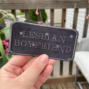 May include: A rectangular, dark grey patch with the words "LESBIAN BOYFRIEND" embroidered in light grey thread. The patch has a stitched border and star accents. The patch is held in a person's hand.