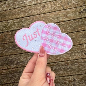 May include: Two heart-shaped patches. One is light blue with the word "Just" embroidered in pink, and the other is pink gingham with the words "Robbed a House" embroidered in white. Rhinestones are on the light blue patch.