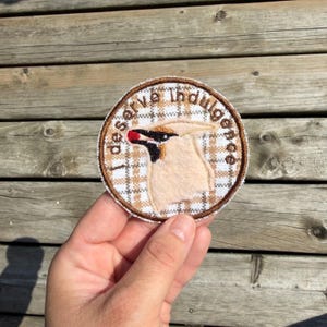 May include: A circular embroidered patch featuring a beige bird with a black and red beak. The background is a white and tan plaid pattern. The text "I deserve indulgence" is stitched in brown letters along the upper edge of the circular border.