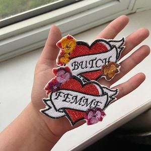 May include: Two embroidered iron-on patches in the shape of hearts with floral accents. The top patch says "Butch" and the bottom patch says "Femme".