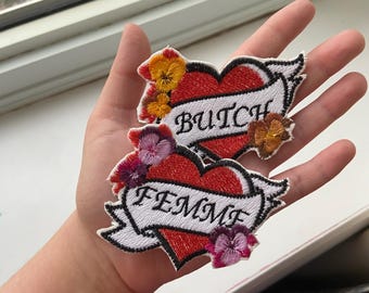 Może przedstawiać: Dwa haftowane naszywki w kształcie serca z kwiatowymi akcentami. Górna naszywka głosi "Butch", a dolna "Femme".