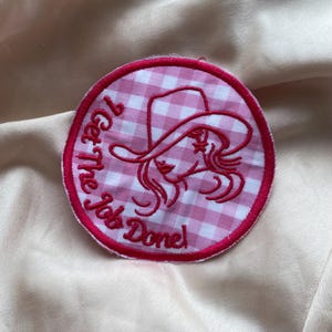 Può includere: Toppa in tessuto vichy rosa e bianco con contorno ricamato rosso. La toppa presenta una donna dei cartoni animati con un cappello da cowboy e il testo "I Get the Job Done!"