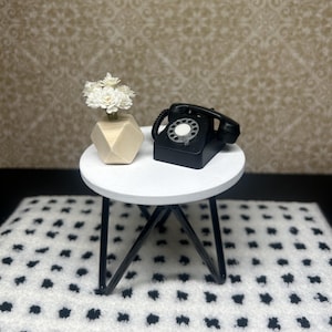 May include: A black rotary phone and a white geometric vase with white flowers sit on a white round table with a black metal tripod base. The table is on a white rug with black polka dots.