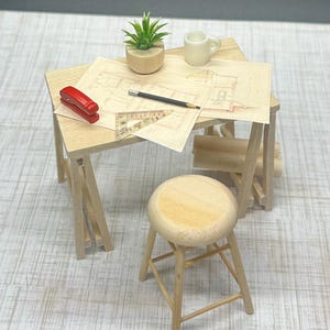 Könnte beinhalten: Miniatur-Architektentisch und Hocker aus Holz. Auf dem Tisch befinden sich ein roter Tacker, ein Bleistift und Baupläne. Eine kleine Pflanze und eine weiße Tasse sind ebenfalls vorhanden. Der Hocker ist rund und hat vier Beine, alles aus hellem Holz.