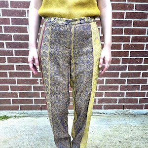 May include: High-waisted, patterned pants with a button closure. The pants feature a mix of gold, navy, and burgundy colors. The design includes a floral pattern and vertical stripes. The model is wearing a mustard yellow sweater.