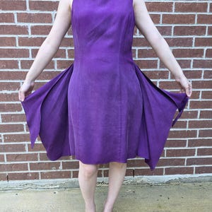 May include: A sleeveless, knee-length purple dress with a fitted bodice and flared skirt. The dress is made of a textured fabric and is being held open to display the skirt's design. The background is a red brick wall.