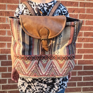 May include: A large, multi-colored woven backpack with a brown faux leather flap and buckle closure. The backpack has a geometric pattern with red, blue, and yellow accents.