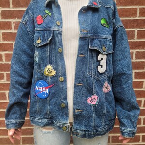 May include: A blue denim jacket with various patches, including a cherry, a heart with the text "Drop Dead", a heart with the text "Go Away", a NASA logo, and a heart with the text "Back Off".