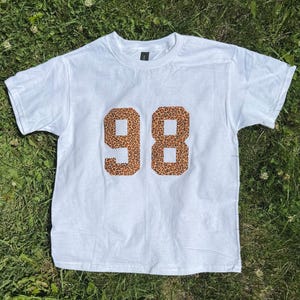 May include: A white t-shirt with the number "98" in a leopard print design. The shirt is short-sleeved and has a crew neck. The shirt is laying on a bed of green grass.
