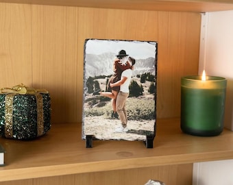 May include: A rectangular slate photo plaque with rough, chiseled edges, displaying a printed image of two people embracing in a mountainous landscape. The plaque is supported by two small black plastic stands on a wooden shelf surface.