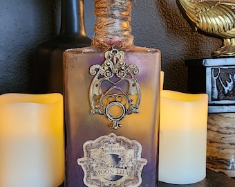May include: A glass bottle with a purple tint and a gold metal design. The bottle has a label that reads "Mystical Apothecary Moon Lily".