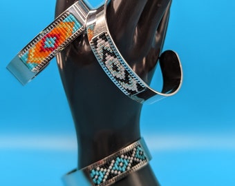 May include: Three silver cuff bracelets with detailed beaded patterns. One bracelet has orange, turquoise, and red beads. Another features a black, white, and turquoise design. The third bracelet displays a turquoise and black pattern.