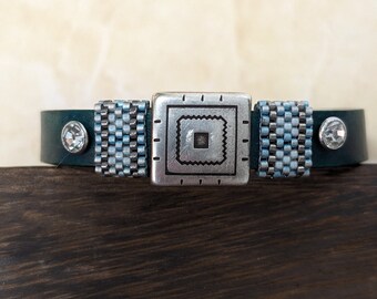 May include: A teal leather bracelet with a silver square centrepiece featuring a geometric design. The bracelet has beaded sections in shades of blue and silver, and is accented with two clear crystal studs.