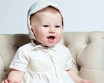 May include: A baby wearing a white bonnet and a white, smocked, short-sleeved onesie sits on a light beige chair. The baby is smiling and looking to the right.
